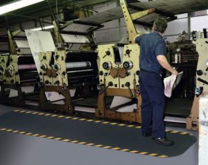 Man standing on a 5/8" thick Hog Heaven Industrial anti fatigue mat runner with safety yellow borders in front of an industrial printing press